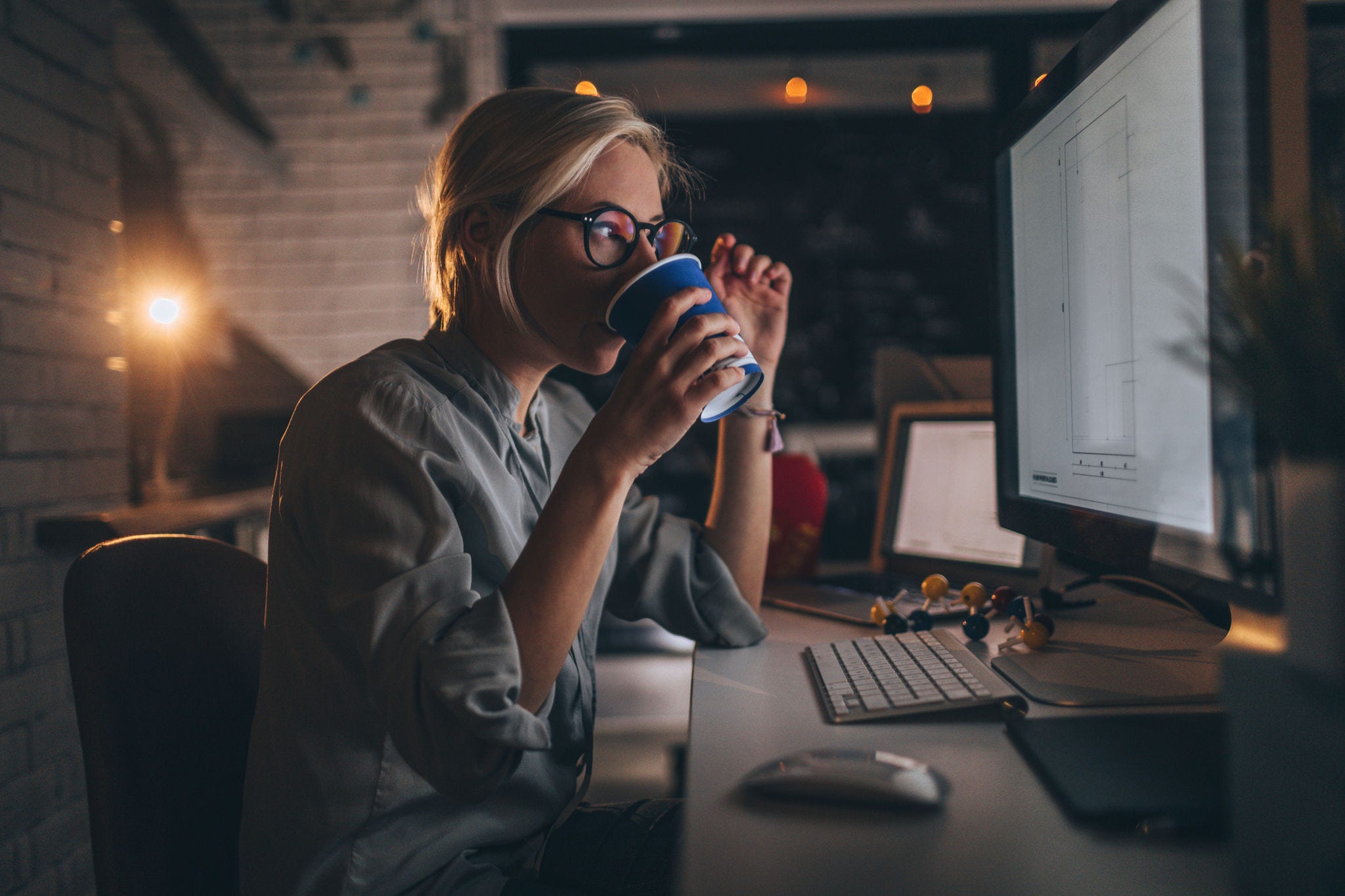 A young woman with blonde hair and glasses is seated at a desk in a dimly lit office, holding a coffee mug while gazing at a large computer screen displaying architectural plans. She appears focused and thoughtful as she examines the details on the screen. Her workstation is equipped with multiple screens, a keyboard, and various other office supplies, creating a productive environment. Soft lighting from a desk lamp and ambient lights in the background add a warm atmosphere to the setting.