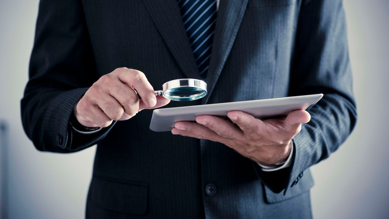 A businessman holding a magnifying glass while looking at a tablet.