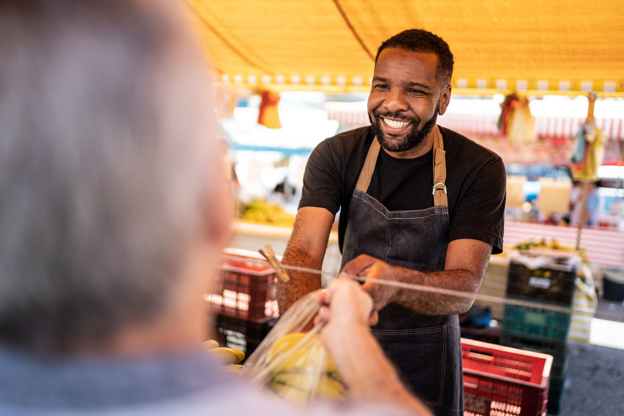 A friendly market vendor in a dark apron smiles warmly while handing a bag of groceries to an out-of-focus customer in the foreground.