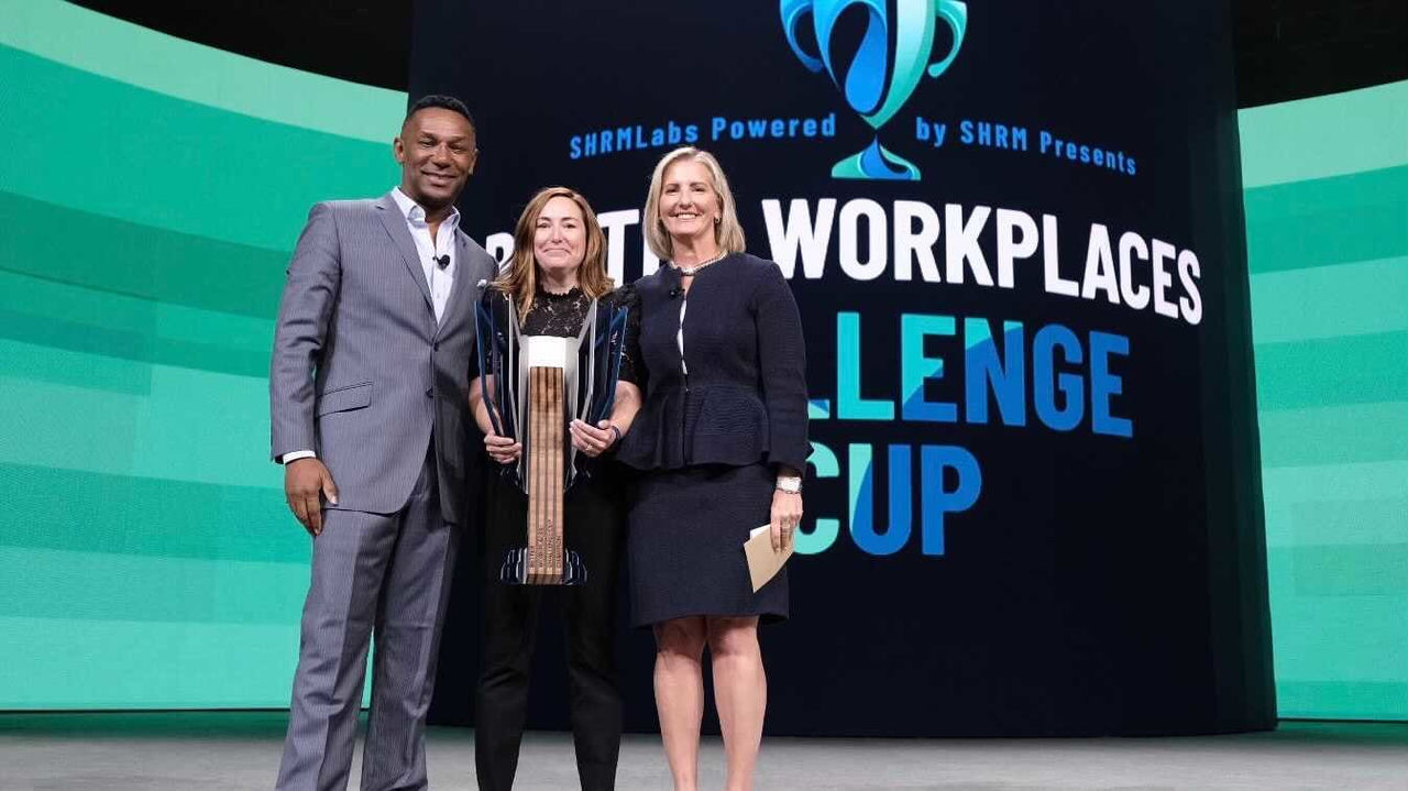 Three people standing in front of a stage with the words workplace challenge cup.