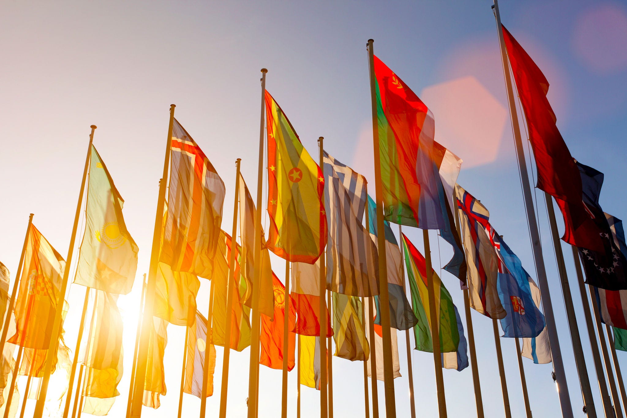497160831 Group of many colorful internationl world flags waving in the sky at sunset, with shining sun and lens flare, low angle view.