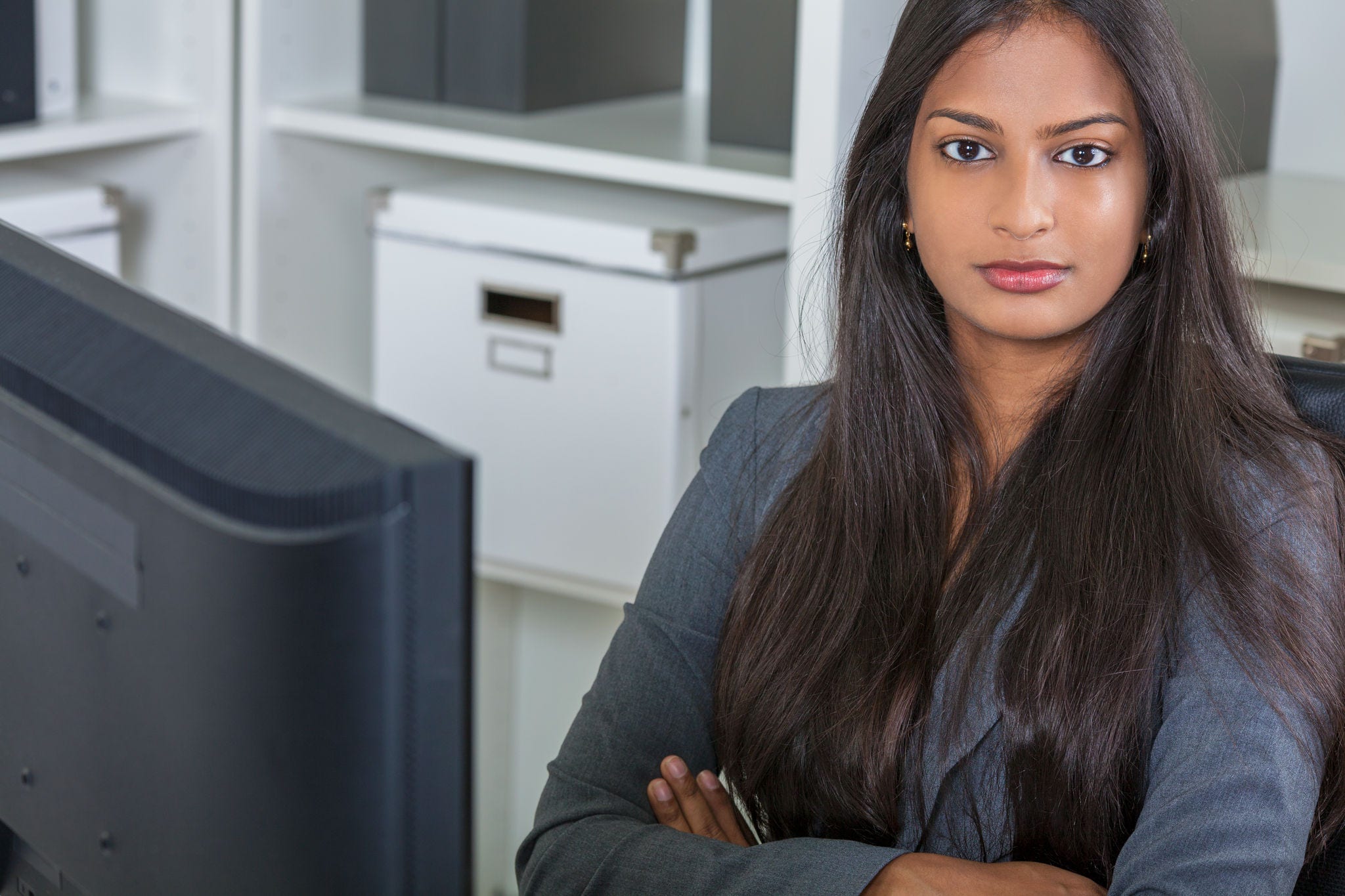 Portrait of a beautiful powerful young Asian Indian woman or businesswoman in office sitting at a desk arms folded with a computer