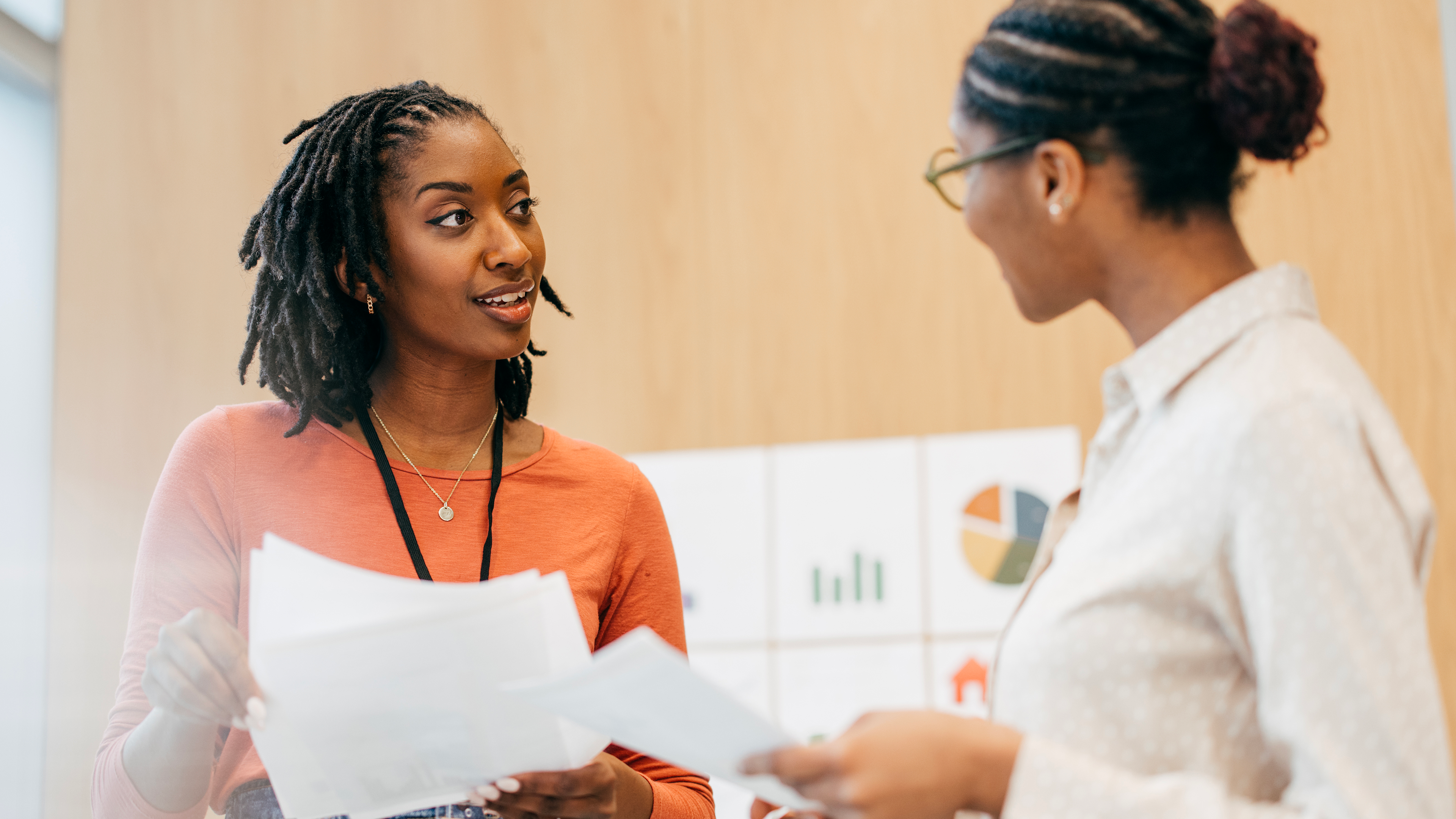 two women speak while holding papers