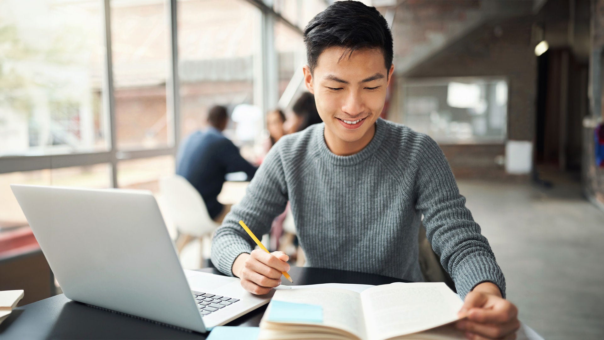 man studying with book and laptop, holding pencil smiling