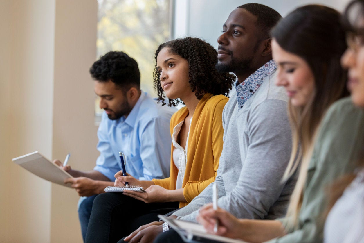 A group of business people sit in a row in a training class.  They look at an unseen speaker as they concentrate on his lecture.