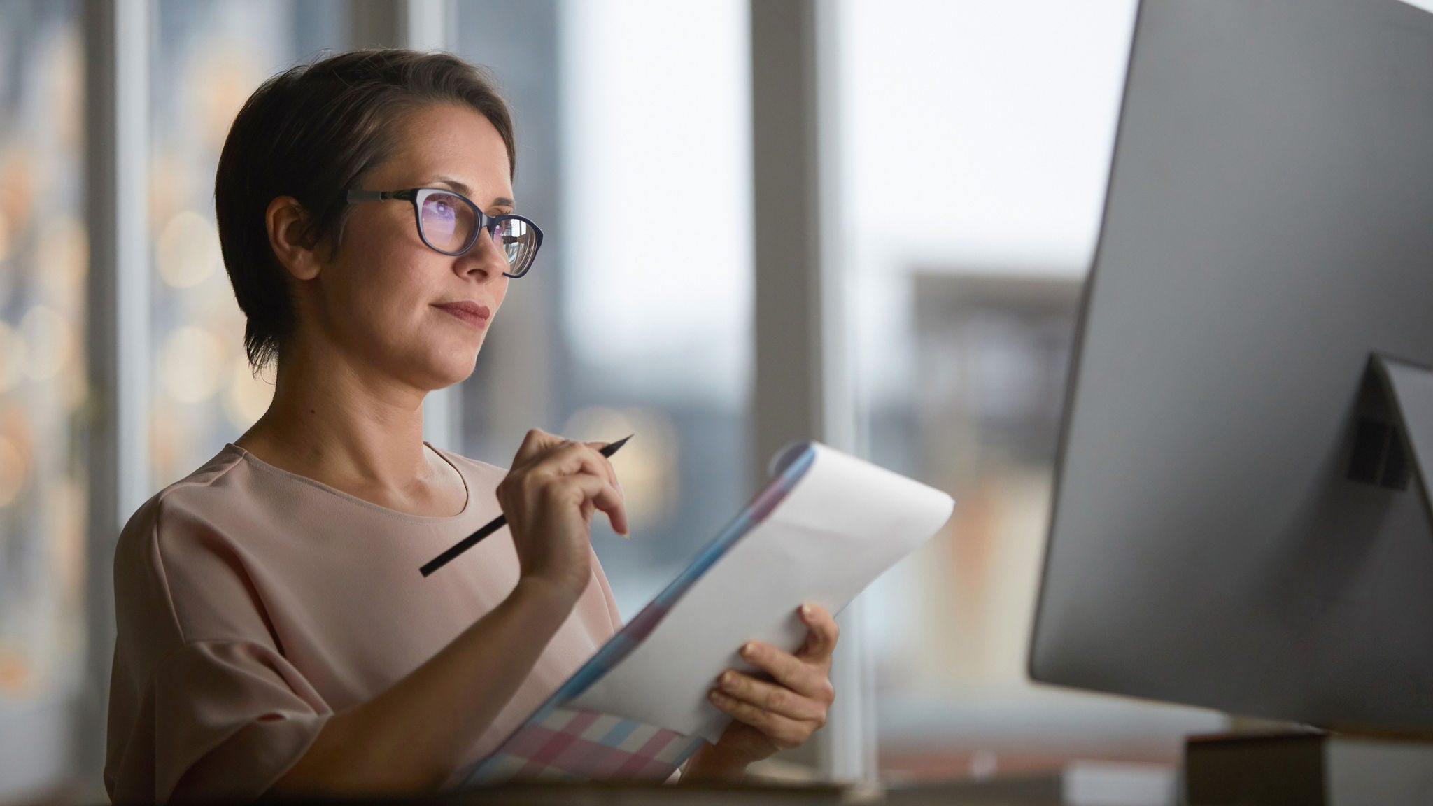 Confident businesswoman with document looking at computer screen and making working notes in office