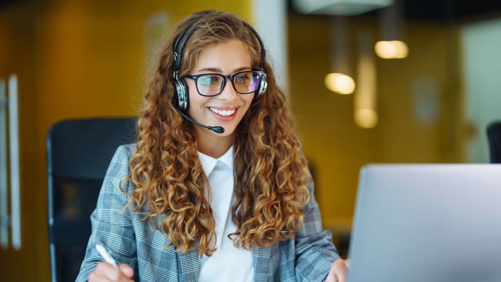 A woman wearing a headset is sitting at a desk in front of a laptop.