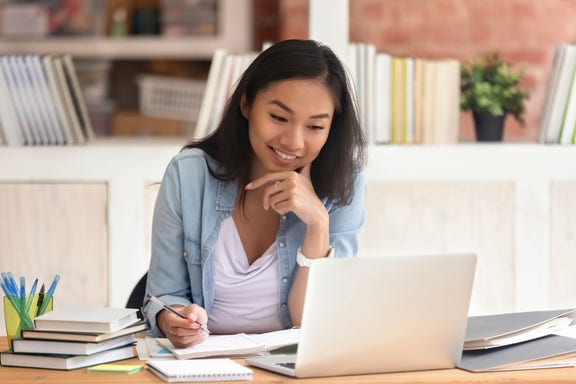 Smiling asian student studying in library with laptop books doing online research for coursework, <a href=