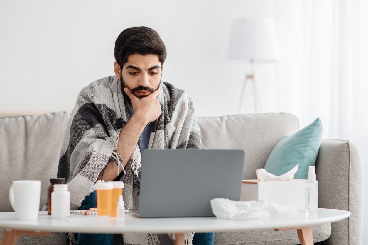 Sick arab male employee suffering from virus but need to work, working on laptop computer and taking pills for flu. Guy sitting on sofa at home interior and using pc, copy space