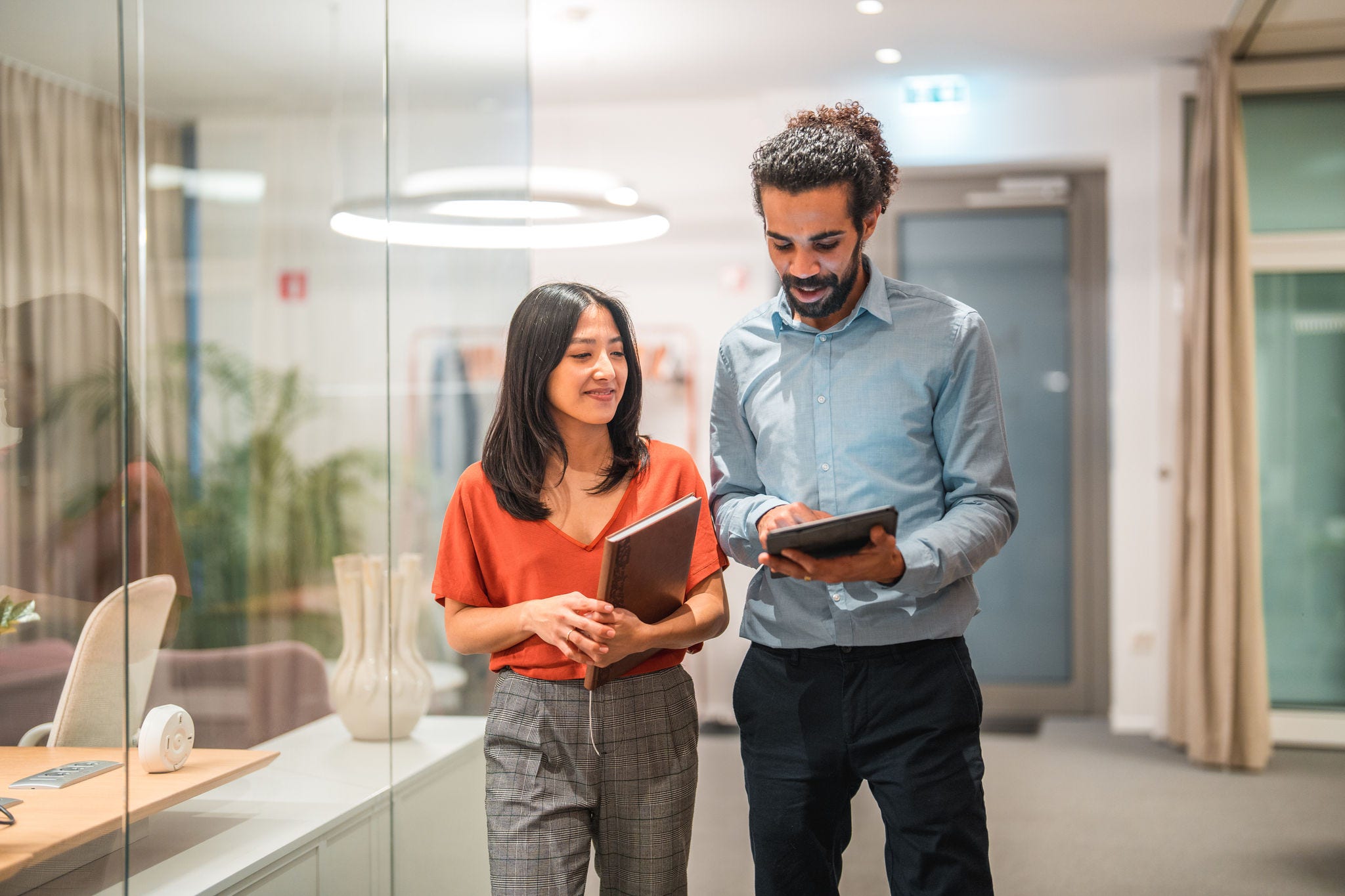 Two handsome, diverse colleagues are seen engaging in a productive conversation while utilizing wireless technology and a digital tablet in a modern office setting. Their smart casual attire adds to their professional appearance as successful employees.