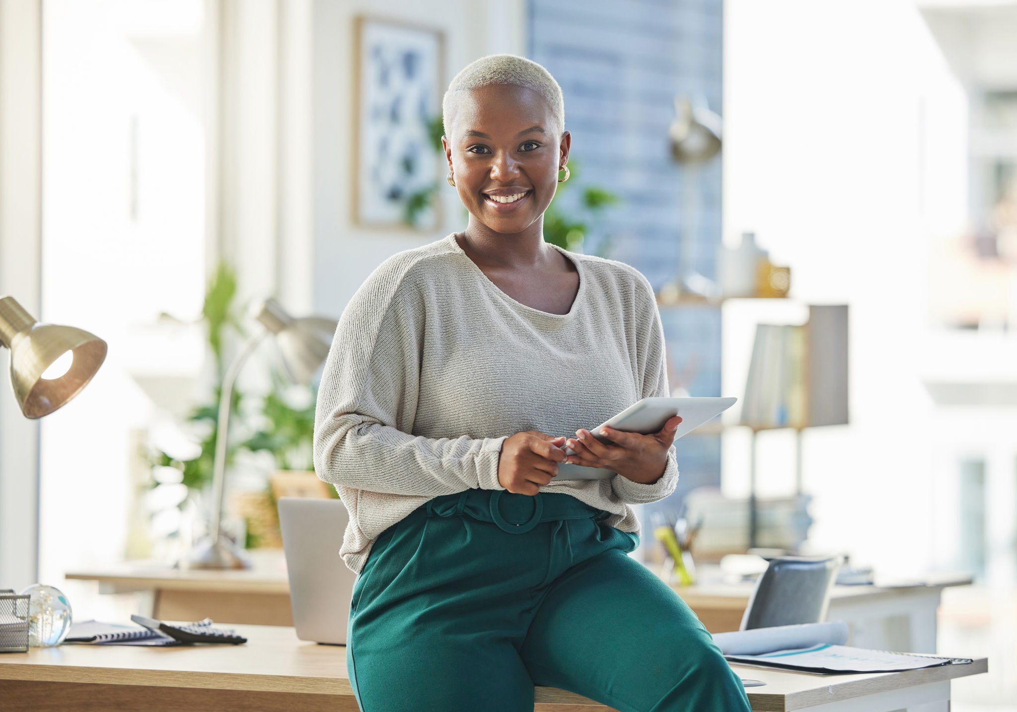 a woman smiles while sitting on a desk holding a tablet computer