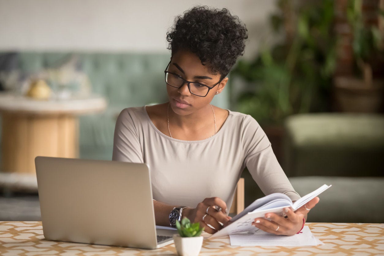 1144287280 Focused young african american businesswoman or student looking at laptop holding book learning, serious black woman working or studying with computer doing research or preparing for exam online