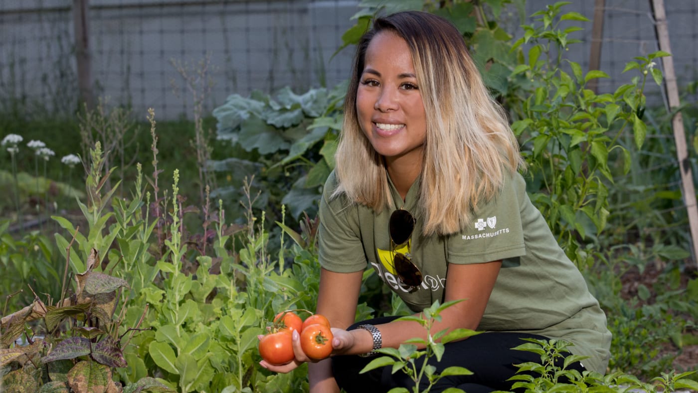 Photo of a smiling woman in a green shirt and sunglasses, kneeling in a community garden filled with vibrant vegetables, holding ripe tomatoes in her hand, representing individual participation in sustainable and green activities.