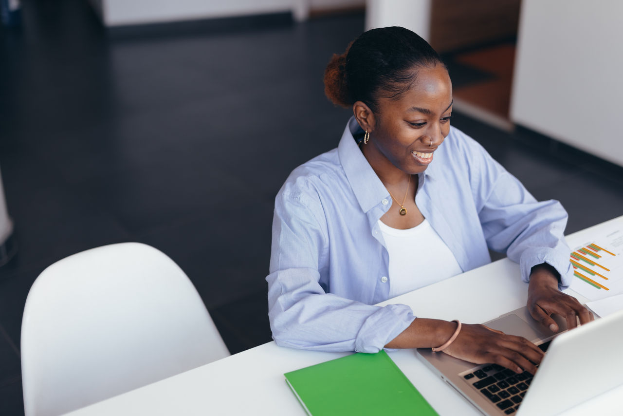 2198392766 woman sitting at a table working on her laptop and smiling