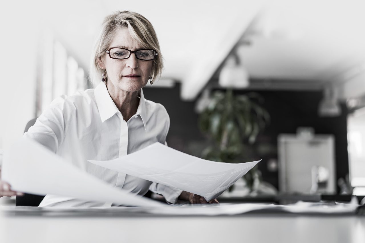 A professional woman examines two pieces of a paper at her desk.