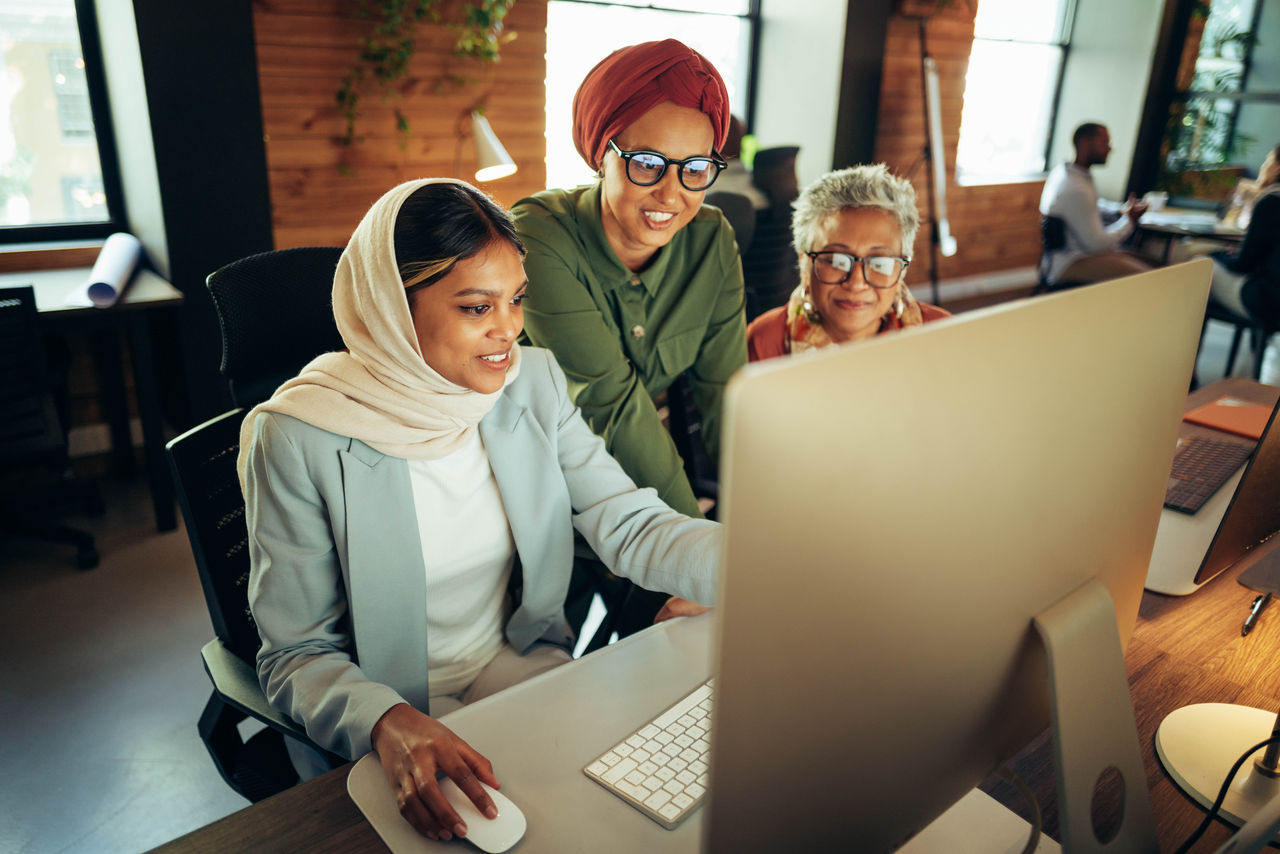 2239873473 Three cheerful businesswomen having a discussion while looking at a computer screen in an office. Multicultural businesswomen working as a team in a modern co-working office.
