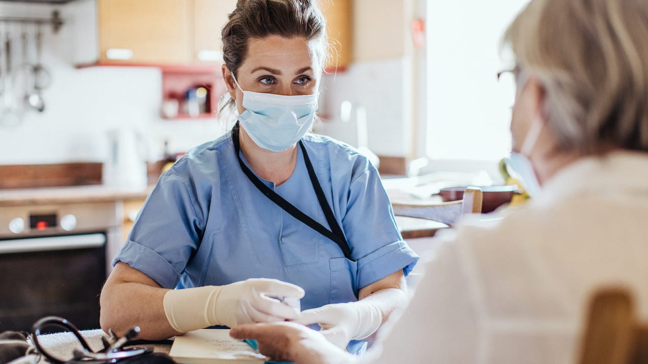 A nurse wearing a mask is helping an elderly woman.