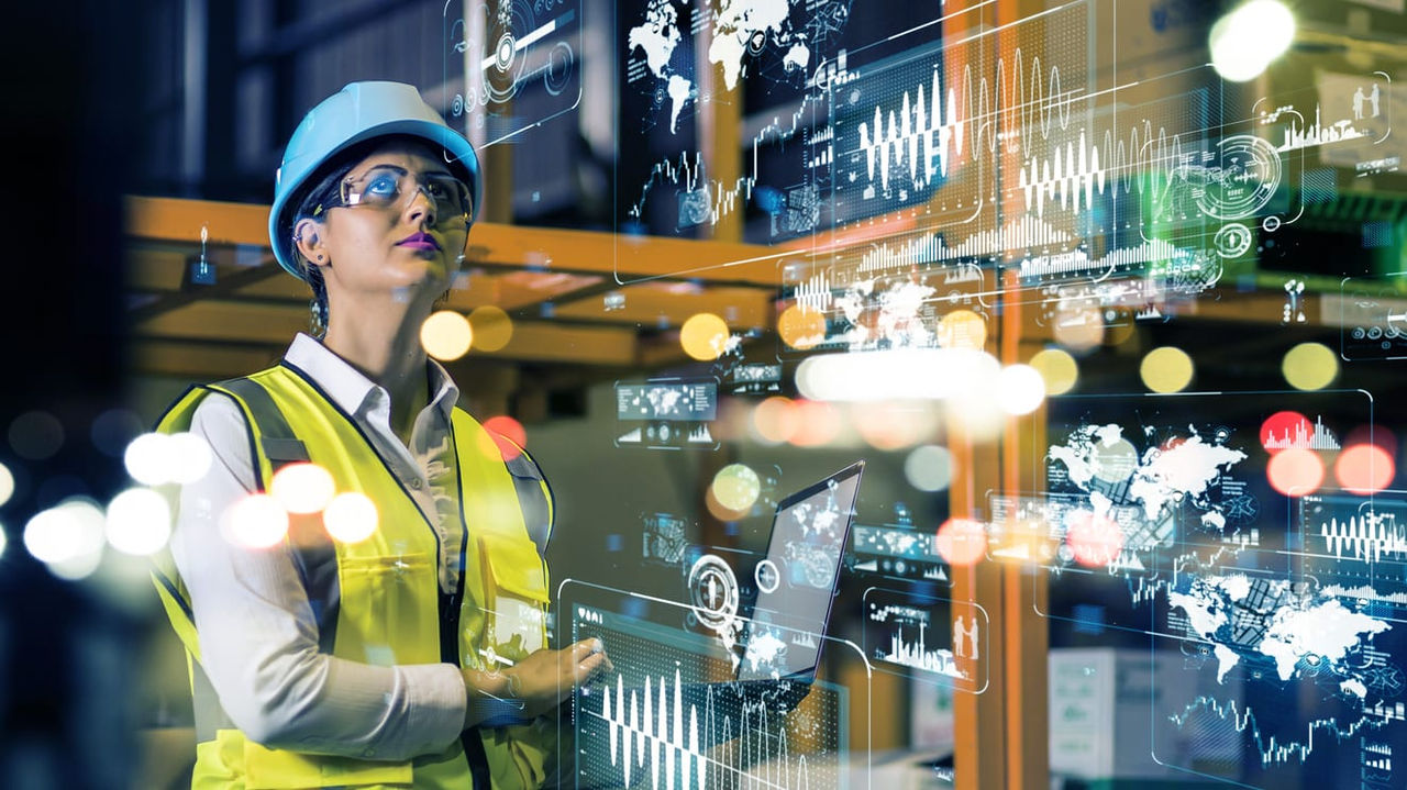 A woman in a safety vest is looking at a computer screen in a warehouse.