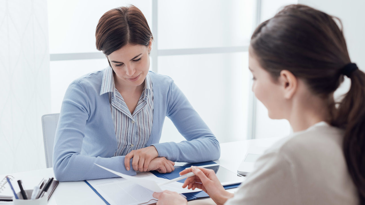 Two women sitting at a desk talking to each other.