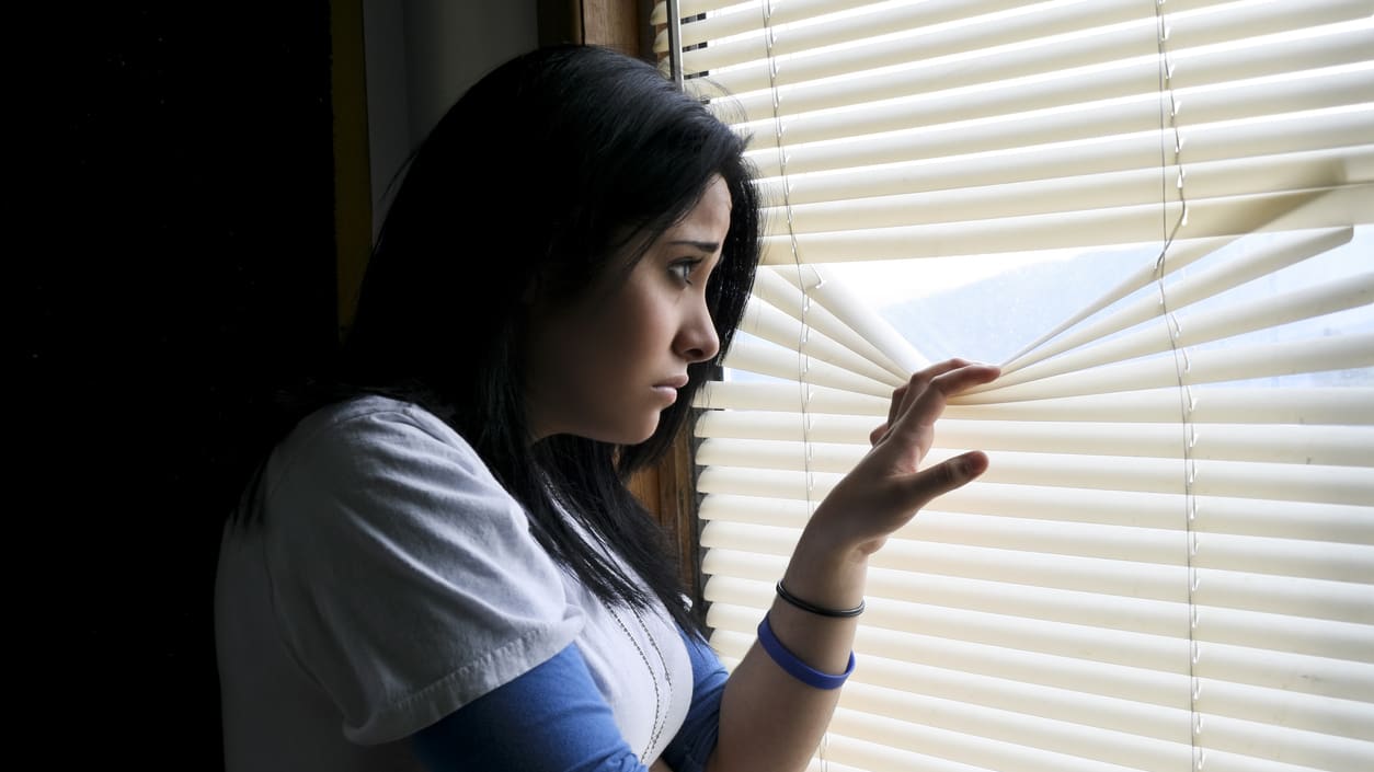 A woman looking out of a window with blinds.