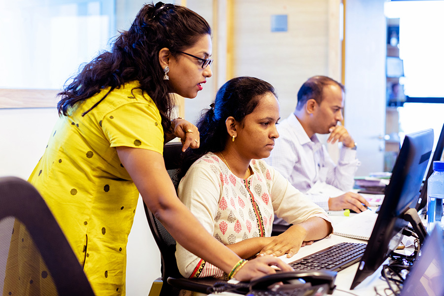 Two employees working at their computers with one getting assistance from a third person.