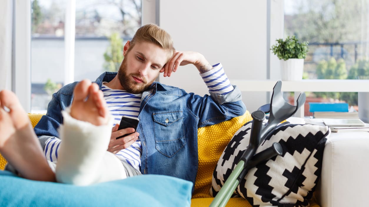 A man sitting on a couch with a broken leg and a cell phone.