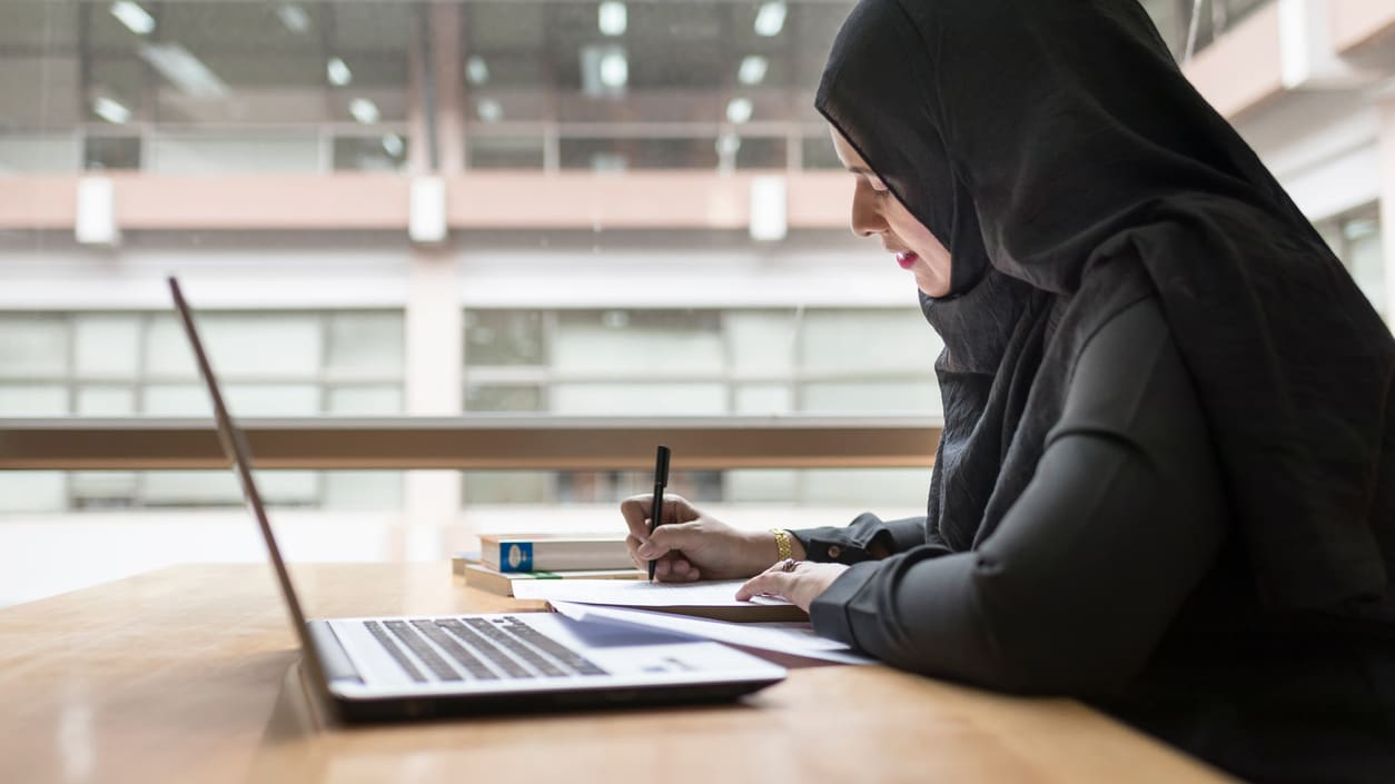 Muslim woman in hijab sitting at a desk with a laptop.