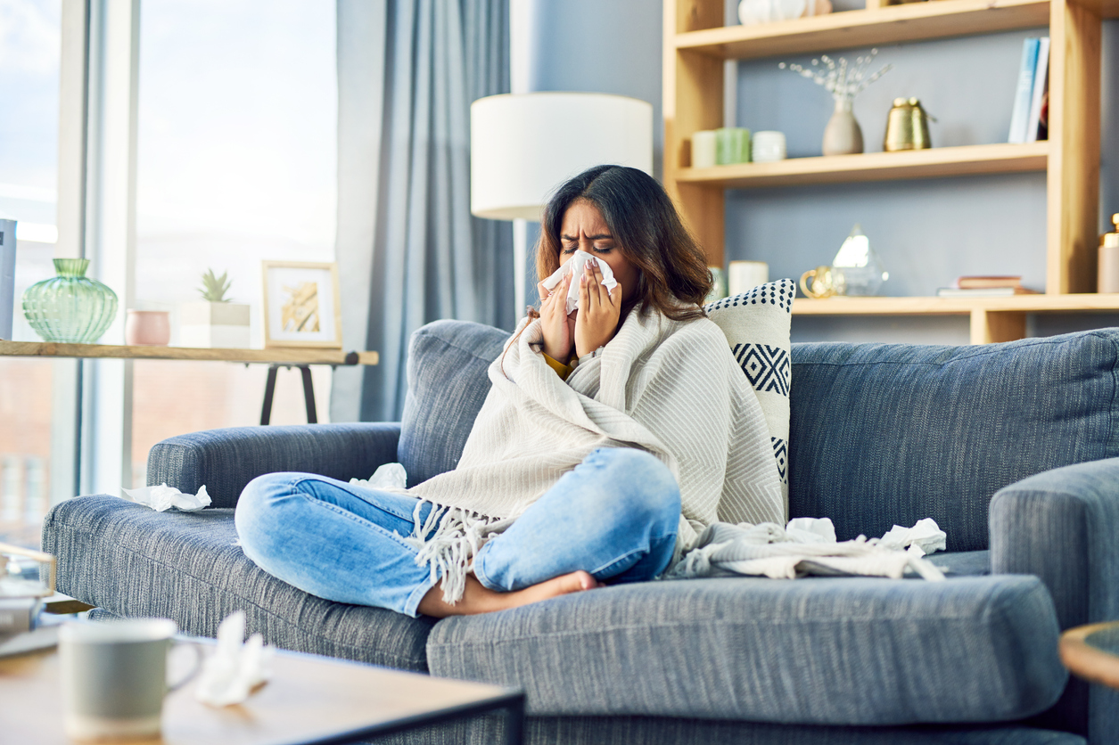 Someone sitting cross-legged on the couch at home and wrapped in a blanket, blowing her nose.