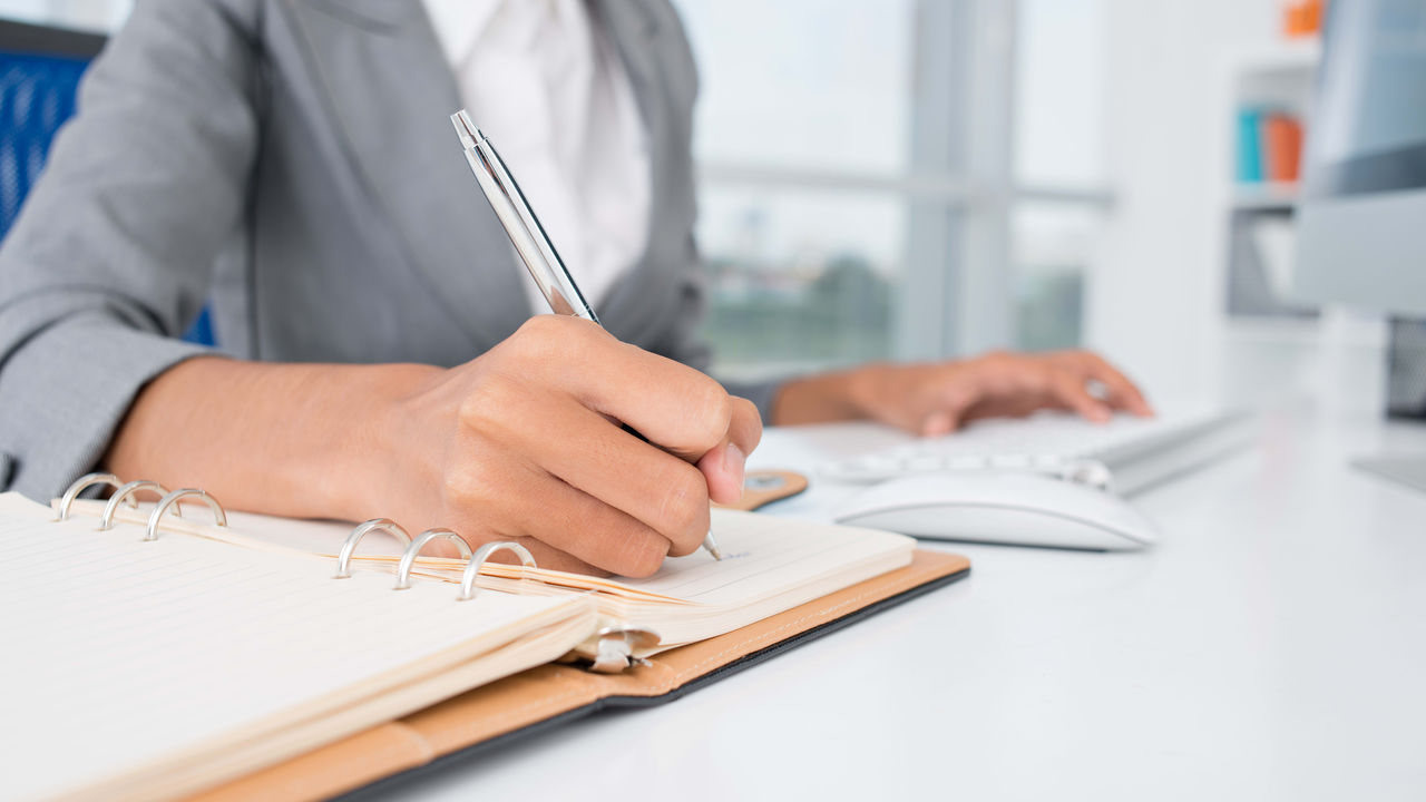 A woman is writing on a notebook in front of a computer.