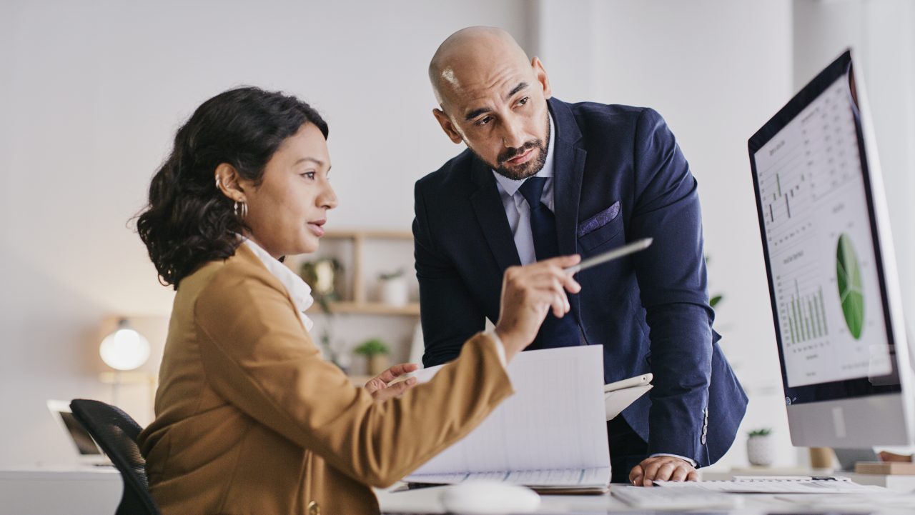 A woman points to a computer screen with budget information on it while a man who's standing looks over her. 
