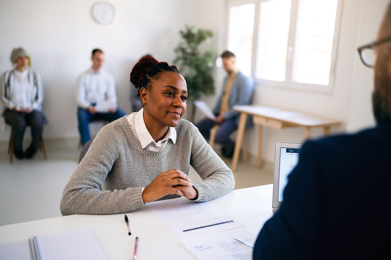 In the office young female candidate of Black ethnicity having a job interview with a male Caucasian recruiter
Other candidates sit on the chair and wait for their turn