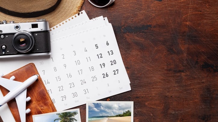 A hat, sunglasses, camera and calendar on a wooden table.