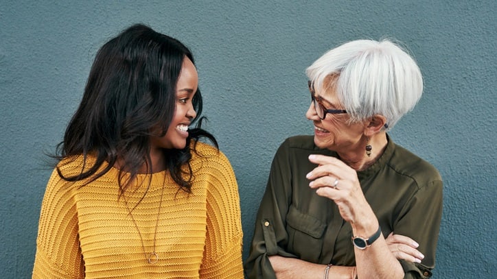 Two women talking against a gray wall.