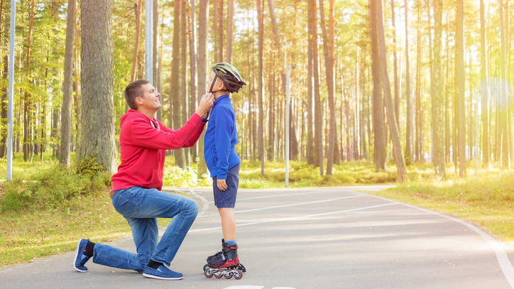 A man is kneeling down to his son on a skateboard in the woods.