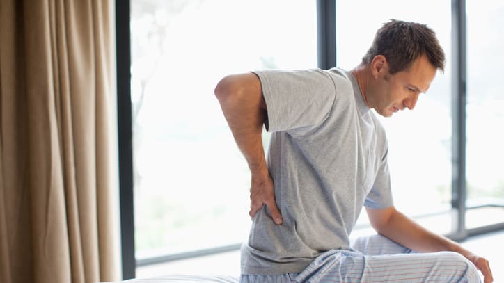 A man in pajamas sitting on a bed touching his back in pain.