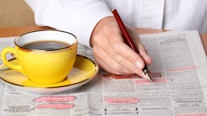 A woman writing on a newspaper with a cup of coffee.