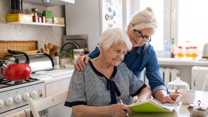 A woman is helping an older woman with a book in the kitchen.