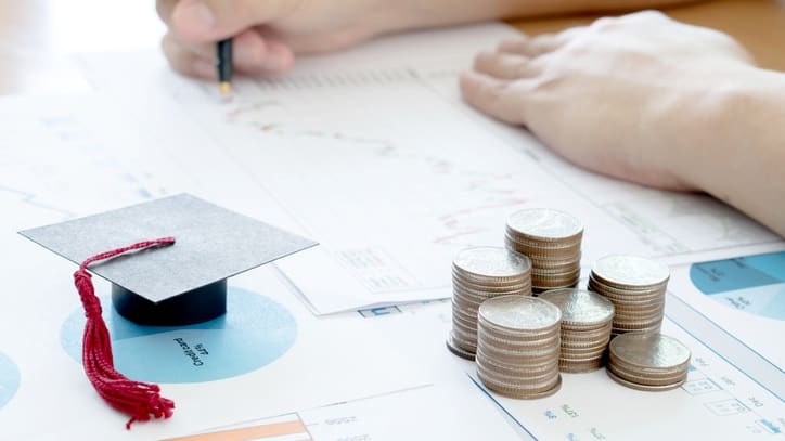 A person writing on a piece of paper with coins and a graduation cap.