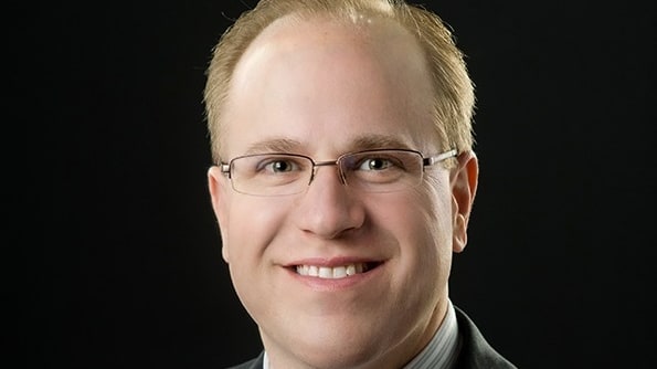 A man in a suit and tie smiles in front of a black background.