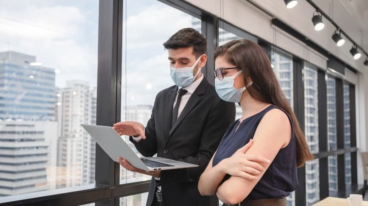 A man and woman wearing face masks are looking at a laptop in an office.