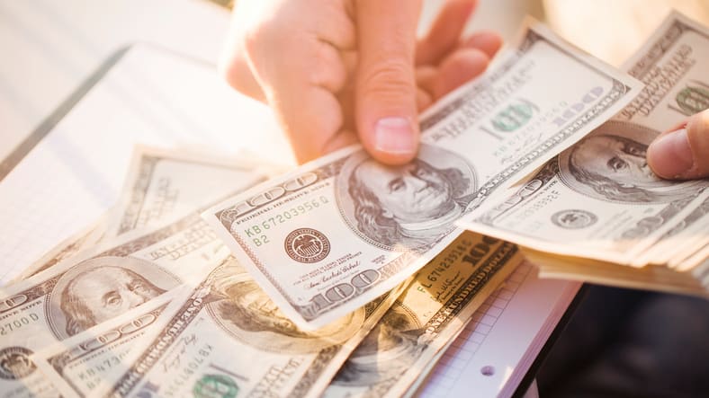 A person is holding a stack of money in front of a laptop.