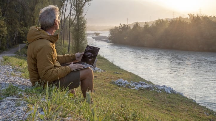 A man is sitting on the side of a river with a laptop.