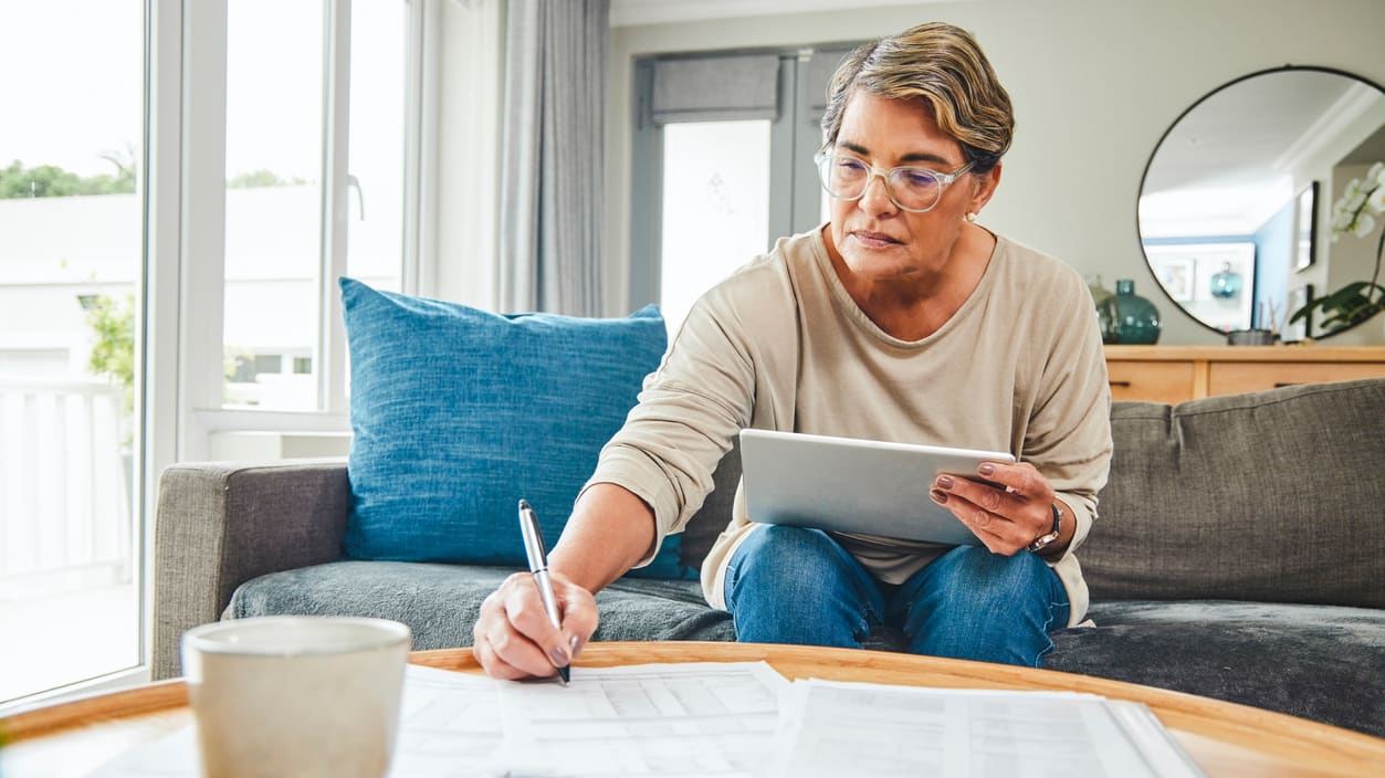 A senior woman is sitting on a couch and writing on a tablet.