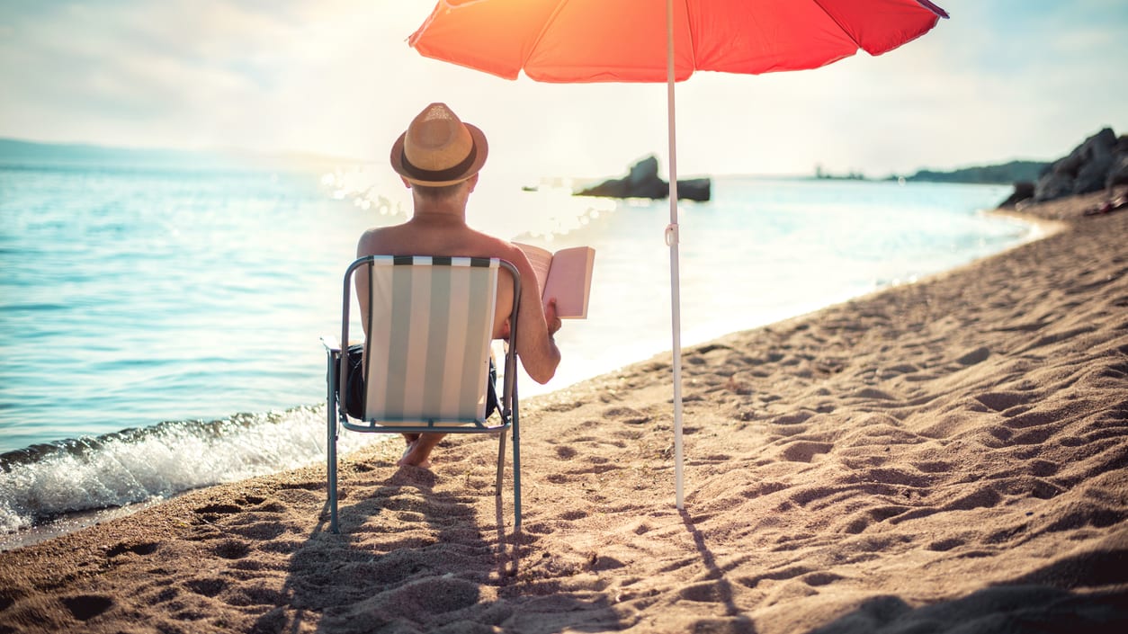 A man sitting in a chair on the beach with a red umbrella.