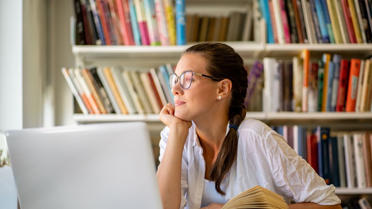 A young woman looking at her laptop in front of bookshelves.