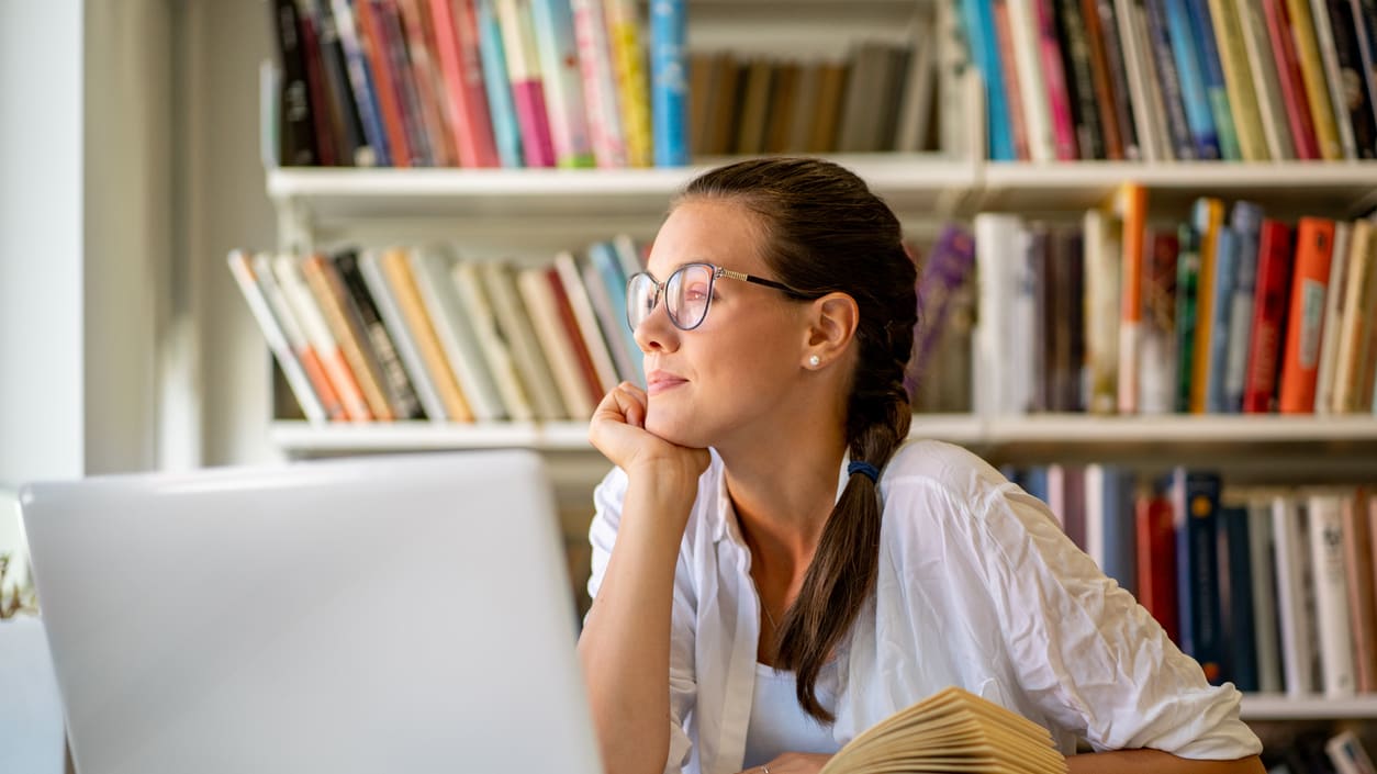 A young woman looking at her laptop in front of bookshelves.