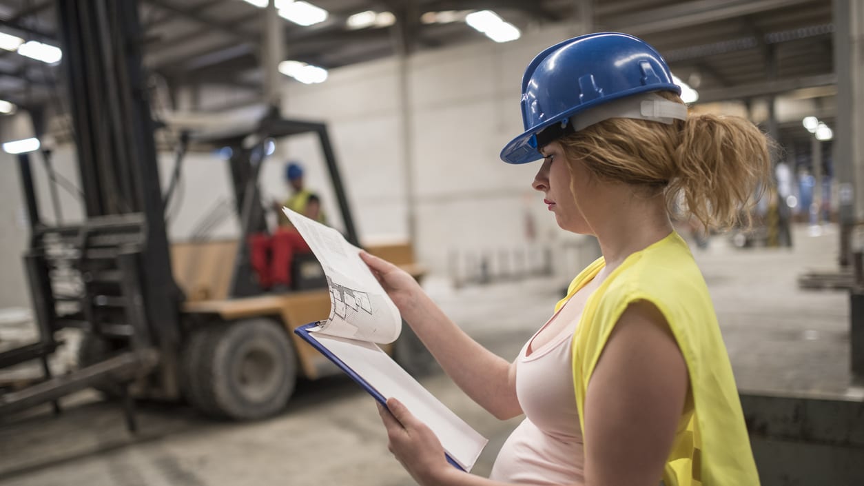 A pregnant woman holding a clipboard in front of a forklift.