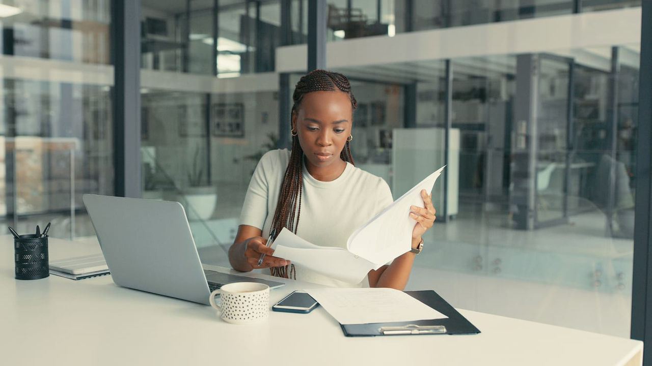 A young african american woman is sitting at a desk with a laptop and papers.