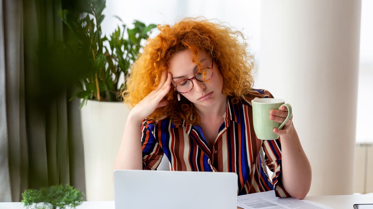 A woman with red hair sitting at a desk with a laptop and a cup of coffee.