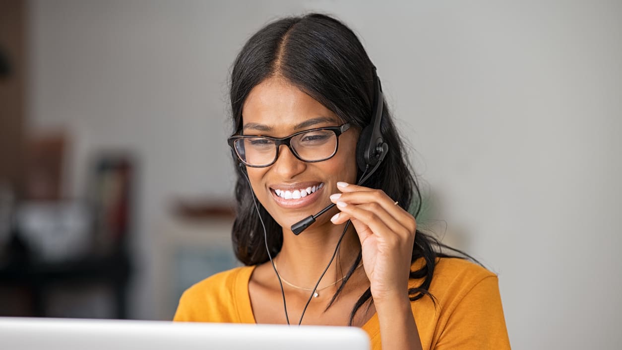 A woman wearing glasses is using a headset while working on a laptop.